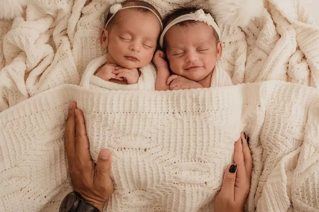 Gêmeas recém-nascidas dormindo juntas sob manta clara no ensaio newborn